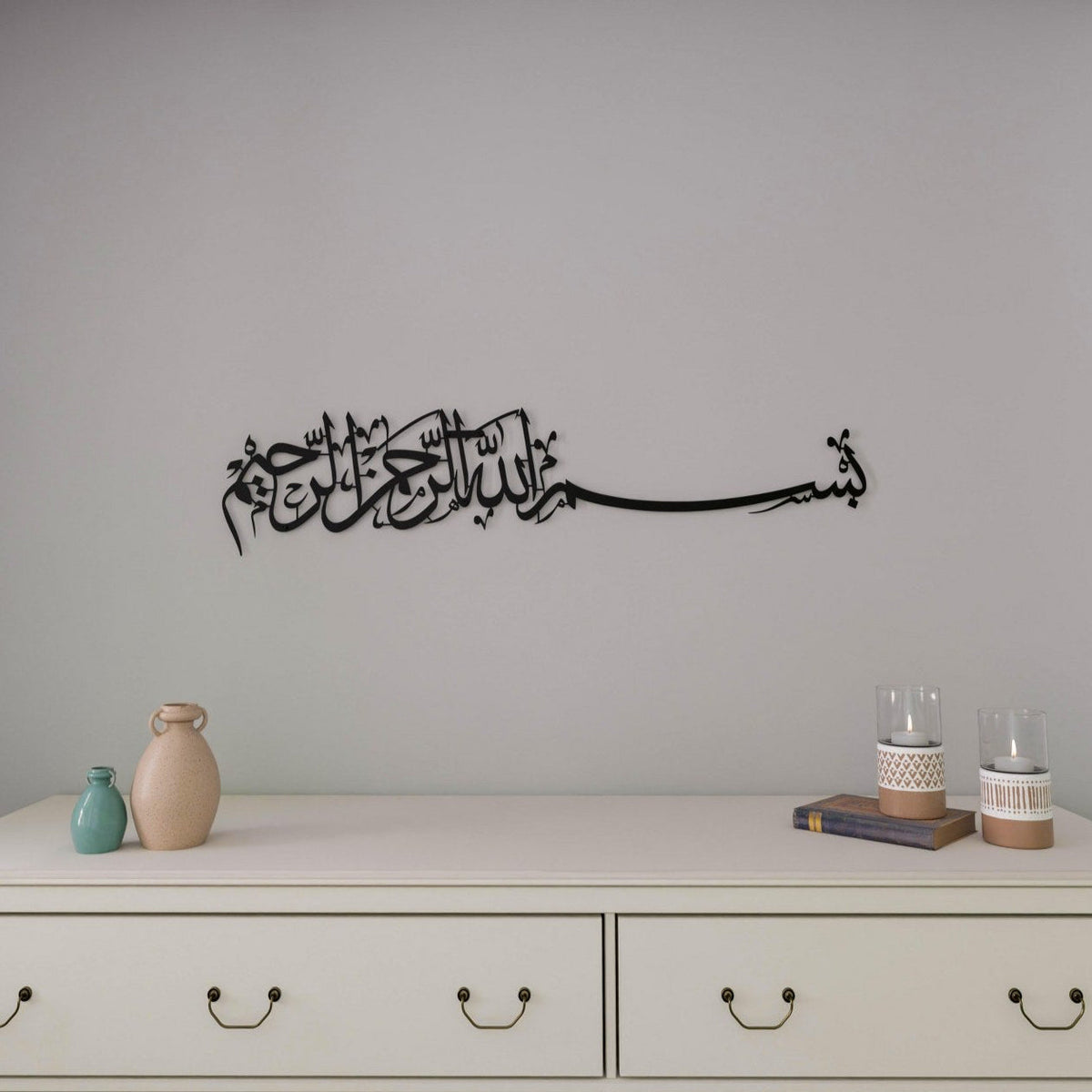 A black cut-metal Bismillah calligraphy band hung on a pale gray wall above a cream chest of drawers with two vases, a book and two candle holders