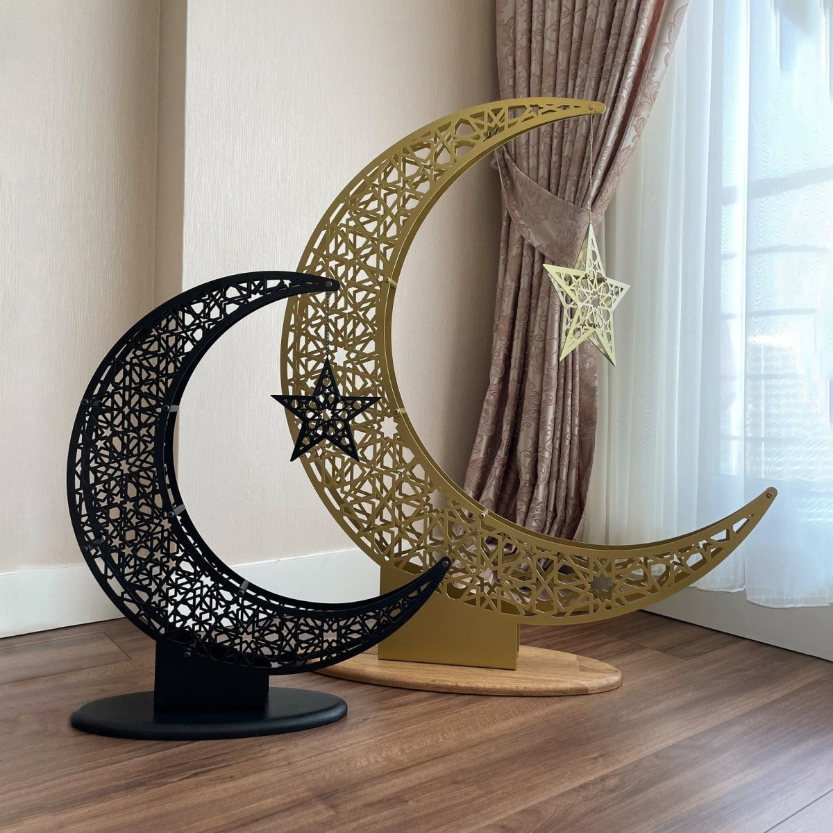 A large gold and a smaller black openwork metal crescent moon, each with a hanging star, standing on round bases beside a curtained window