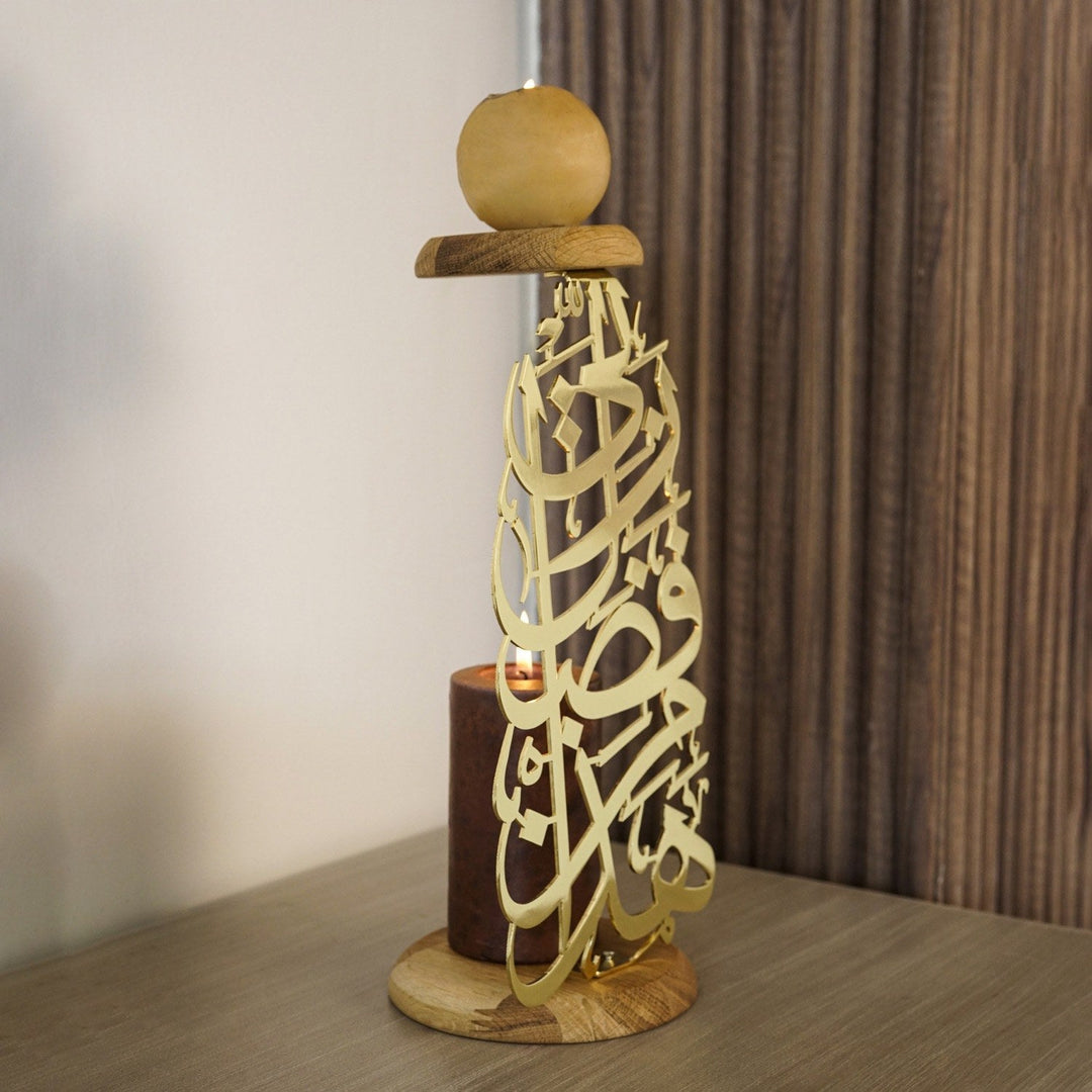 A tall shiny gold candleholder made of interlocking Arabic calligraphy on a round wooden base, with a pillar candle inside, on a table against a curtain