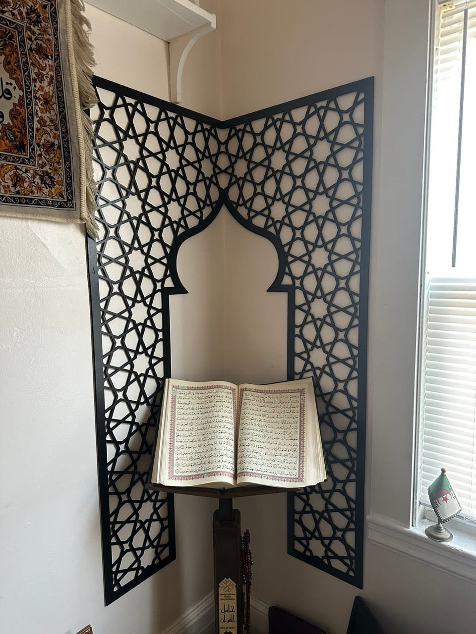 Two black geometric mihrab panels meeting at an internal room corner to form a single arch across the join, with an open Qur'an on a stand in front