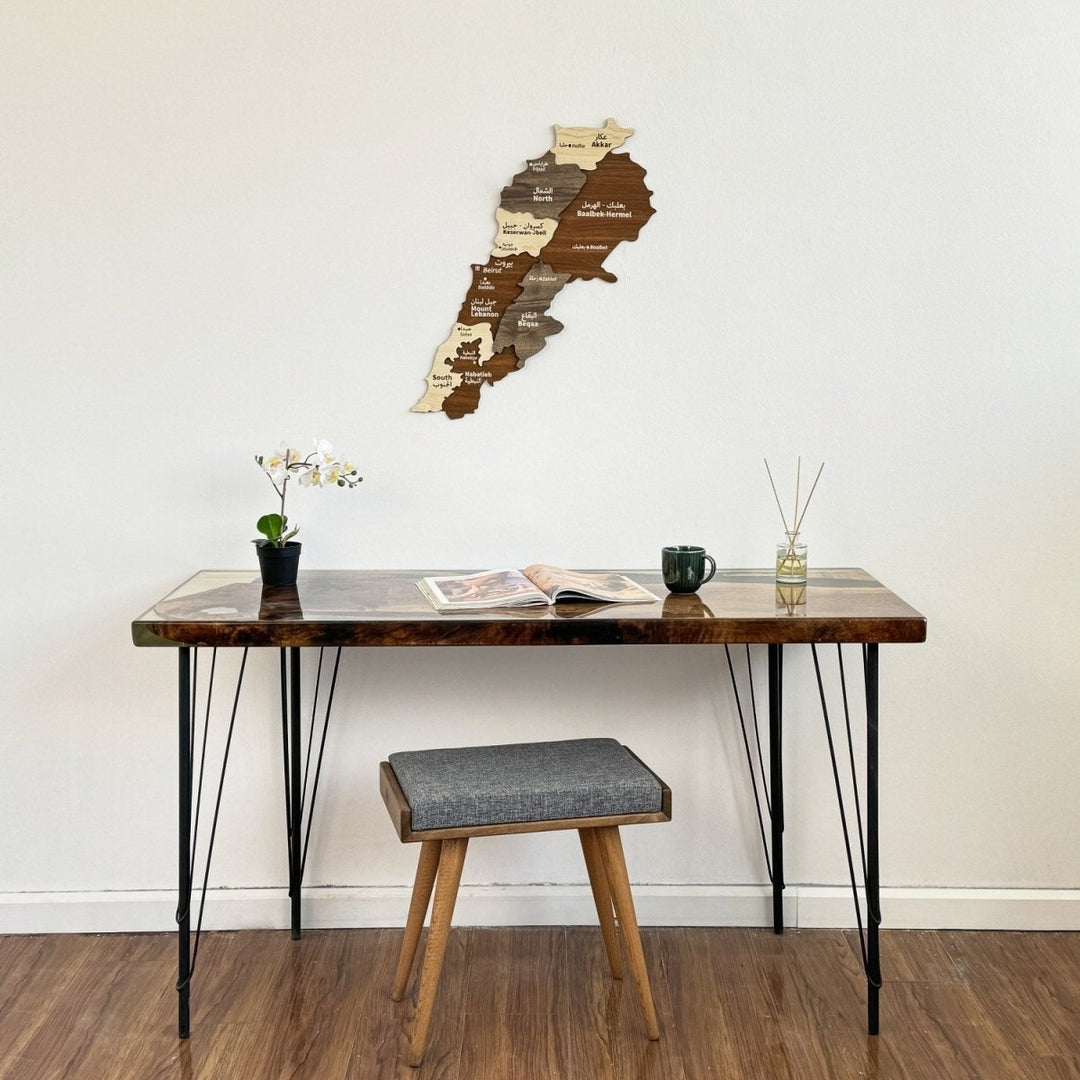 A layered wooden map of Lebanon in walnut, gray and cream tones on a white wall above a walnut-and-resin desk with hairpin legs, an orchid, an open magazine and a gray upholstered stool