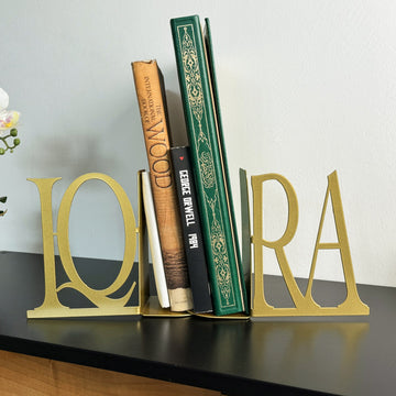 Matte gold IQRA Latin letter bookends holding books on a black cabinet beside an orchid