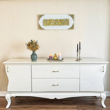 Kalima-Shahada-Glaskunst in goldenem Acryl auf einfarbig weißem Glas über einem weißen Sideboard mit Kerzenständern, weite Ansicht