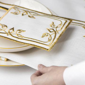 Gold embroidered interlocking scroll napkin laid over a plate on the white placemat, hand at the edge