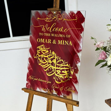 Arabic wedding sign in gold and red on glass, angled view on an easel beside pink flowers