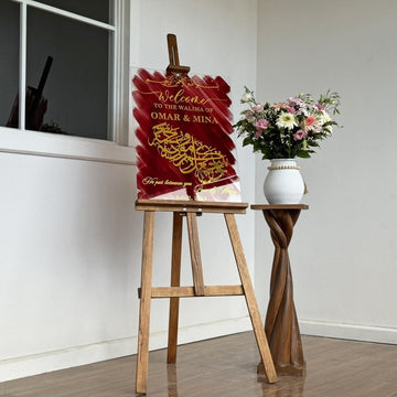 Wedding sign with gold Surah Ar-Rum calligraphy on red glass, standing on an easel beside a vase of flowers
