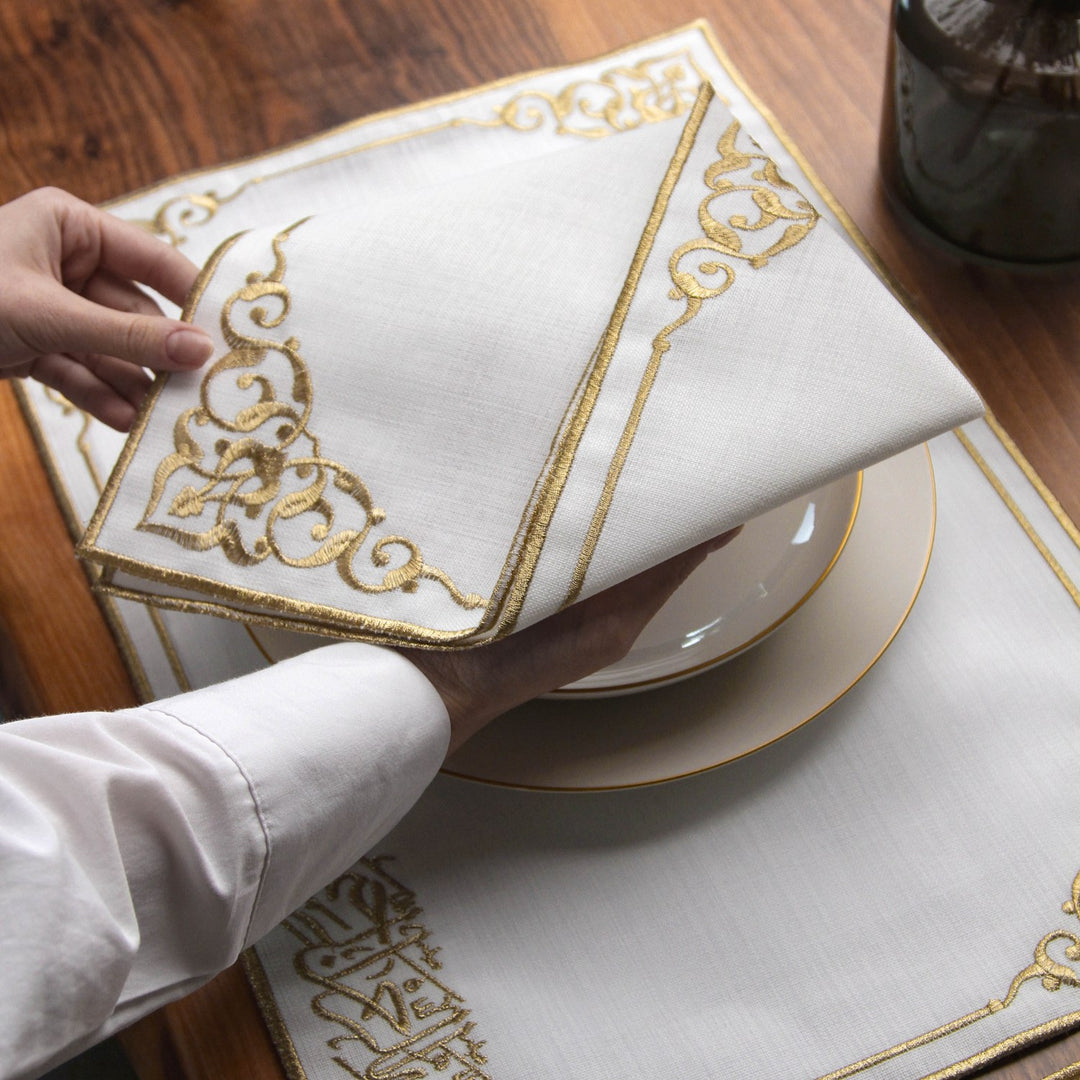 Hand folding a gold embroidered Basmala napkin with ornate corner flourishes over a placemat