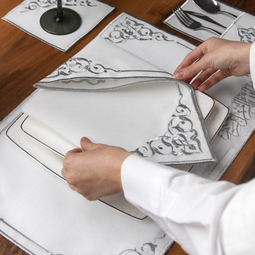 Hand lifting a silver embroidered Basmala napkin to show corner scrollwork over a placemat