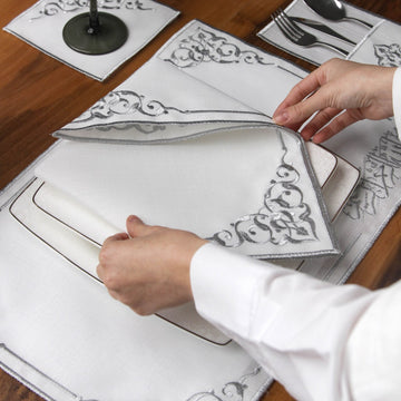 Hand lifting a silver embroidered Basmala napkin to show corner scrollwork over a placemat