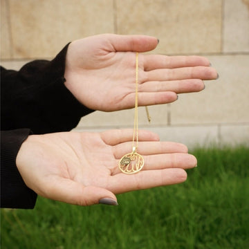 Circular gold Allah necklace resting between two open palms outdoors, showing its size