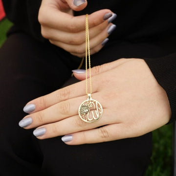 Circular gold Allah necklace dangling from a hand above a ring finger to show its scale