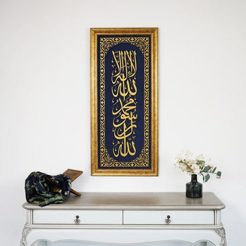 خط كلمة التوحيد الأولى الذهبي المؤطر على مخمل أزرق على جدار أبيض فوق طاولة كونسول