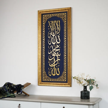 خط كلمة التوحيد الأولى الذهبي المؤطر على مخمل أزرق فوق كونسول أبيض عليه مزهرية ورد