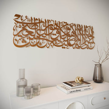 Copper-colored laser-cut Arabic calligraphy wall art mounted on a white wall above a white console table with decorative glass vases and books.