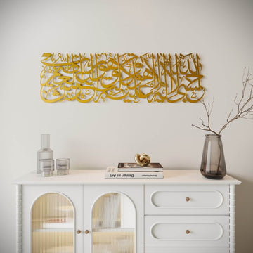 Gold metal Islamic wall art with Arabic calligraphy, displayed above a white console cabinet in a styled interior setting.