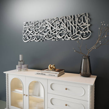 White Arabic calligraphy wall art mounted on a dark gray wall above a white cabinet, styled with books, candles, and a vase in a home interior setting.