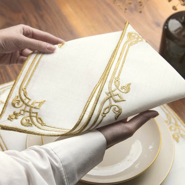 Hands lifting the Gold interlocking scroll napkin to show its double-line embroidered border