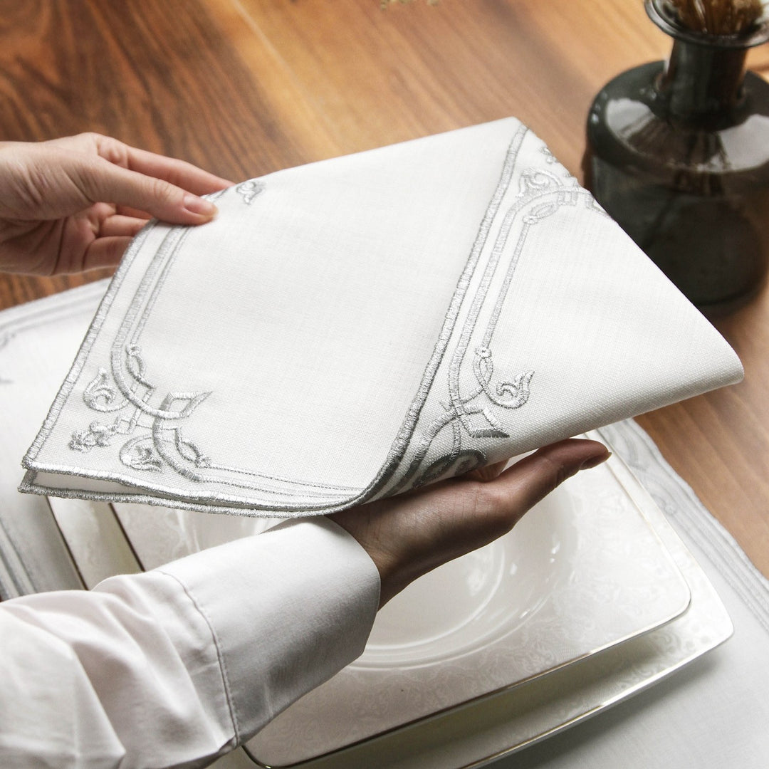 Hands lifting the Silver interlocking scroll napkin over a white plate to show its embroidered border