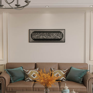 Shahada – Deuxième Kalima en verre trempé, calligraphie en acrylique argenté, centrée au-dessus d'un canapé marron