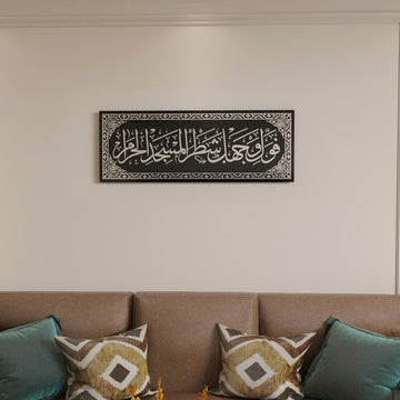 Silver Surah Baqarah 144 mihrab verse glass wall art on black glass, hung above a brown sofa