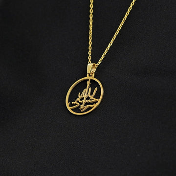 Gold Hasbiyallah pendant tilted on its chain, draped over black fabric
