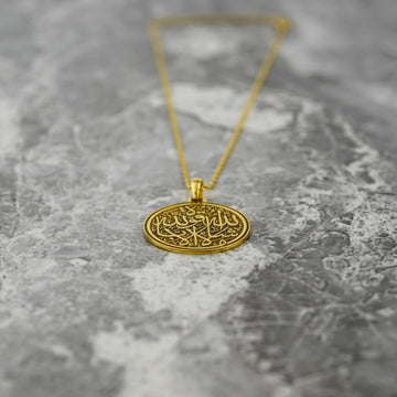 Pendentif disque Mashallah La Quwwata Illa Billah doré posé sur du marbre, la chaîne derrière