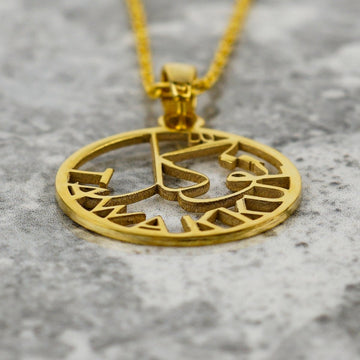 Close-up of the gold Tawakkul pendant lying on marble, showing the cut-out calligraphy and lettering