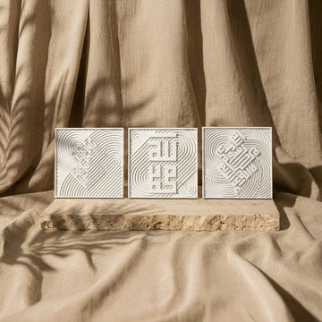 Ensemble de trois carreaux coufiques en relief de pierre blanche en rangée sur une dalle devant des rideaux de lin, vue de face