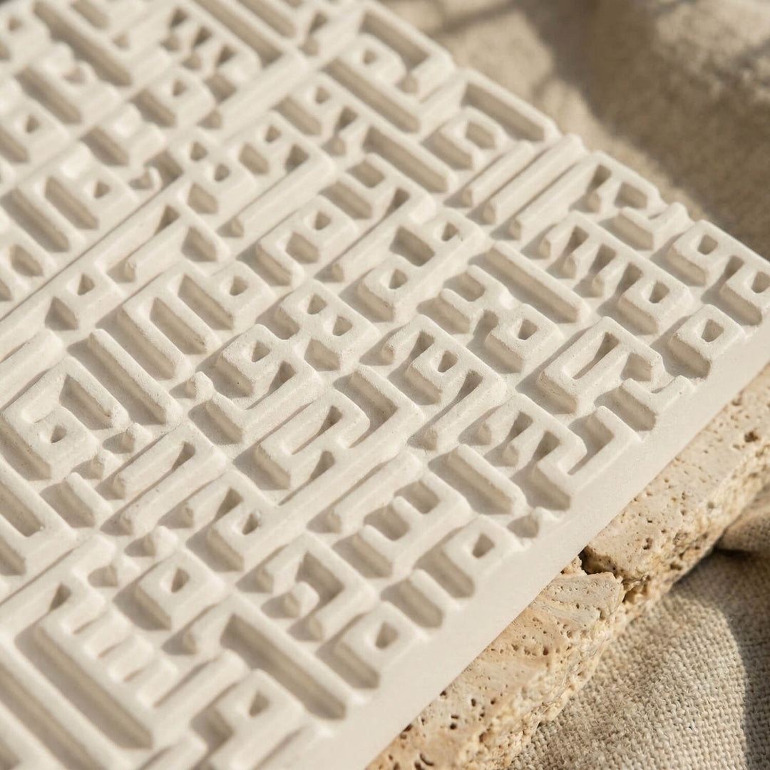 Close-up of raised Kufic calligraphy lines on a white stone relief tile, angled with the tile edge showing