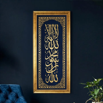 خط كلمة التوحيد الأولى الذهبي المؤطر على مخمل أزرق معلق على جدار كحلي بجانب نبتة