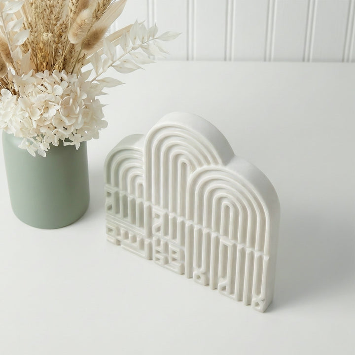 Decorative white arch-shaped object on a white surface with a vase of flowers in the background.