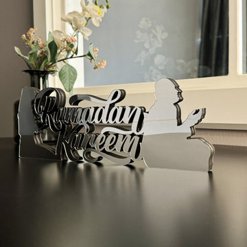 Silver Ramadan Kareem tabletop decor with praying figure in silhouette, front angle by a window