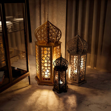 Gold, silver and black Ramadan lanterns glowing together on a marble floor beside curtains