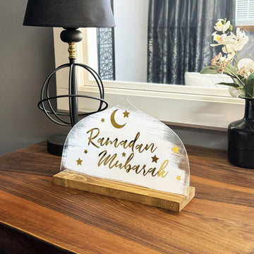 ديكور «رمضان مبارك» صغير بنمط White Strokes على مكتب خشبي بجانب مصباح كروي وورد