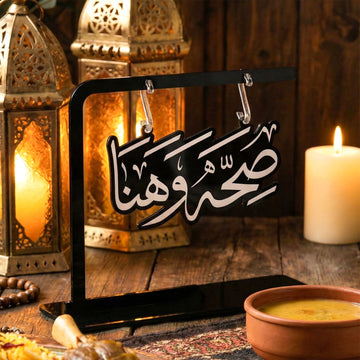 Support Saha w Hana à écriture découpée sur une table d'iftar avec bougie, lanternes et bol de soupe, vue de biais