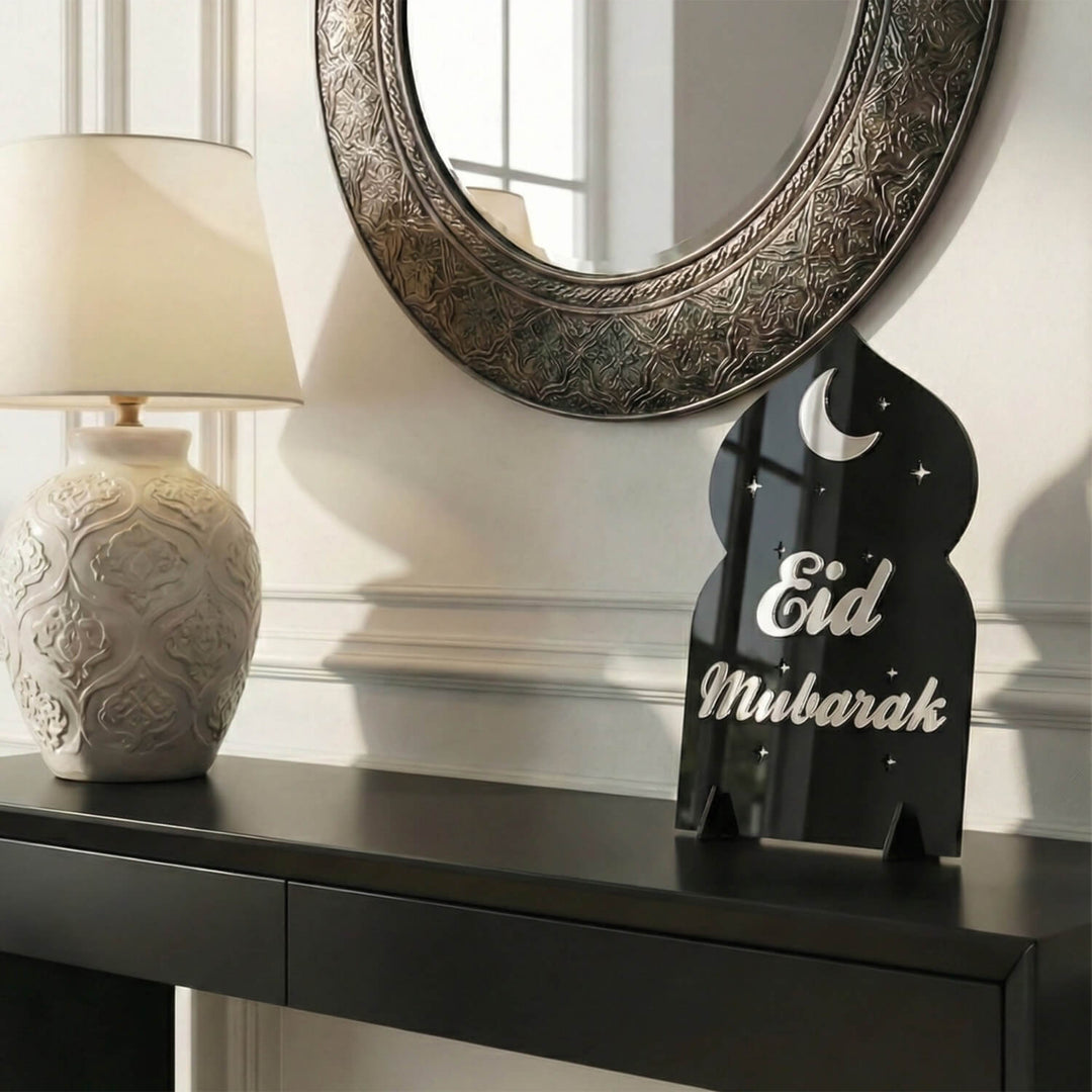 Eid-Mubarak-Acrylaufsteller in Silber auf Schwarz auf einer Konsole neben einer Tischlampe und einem runden Spiegel