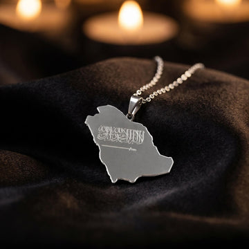 Silver Saudi Arabia map necklace resting on black fabric with candles blurred in the background