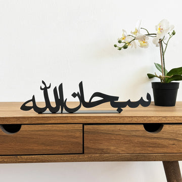 ديكور طاولة معدني أسود «سبحان الله» على مكتب خشبي بجانب زهرة أوركيد