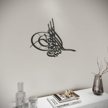 Metall-Wandkunst Tughra Bismillah in Schwarz, an einer hellen Wand, Schrägansicht an einem Buch und einer Vase vorbei