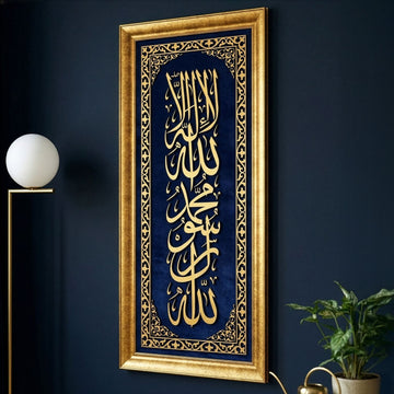 خط كلمة التوحيد الأولى الذهبي العمودي المؤطر على مخمل أزرق بجانب مصباح أرضي على جدار كحلي