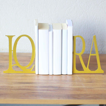 Gold IQRA Latin letter bookends holding white books on a wooden table, front view
