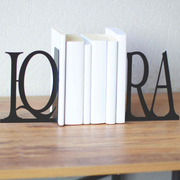 Black IQRA Latin letter bookends holding white books on a wooden table, gray wall behind
