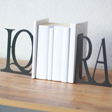 Black IQRA Latin bookends with white books on a wood table, wide front view against a white wall