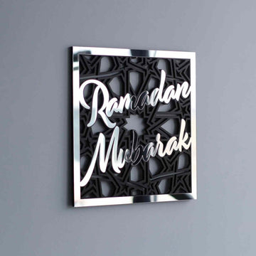 Silver mirror Ramadan Mubarak lettering on a black geometric square wooden wall art, angled on a gray wall